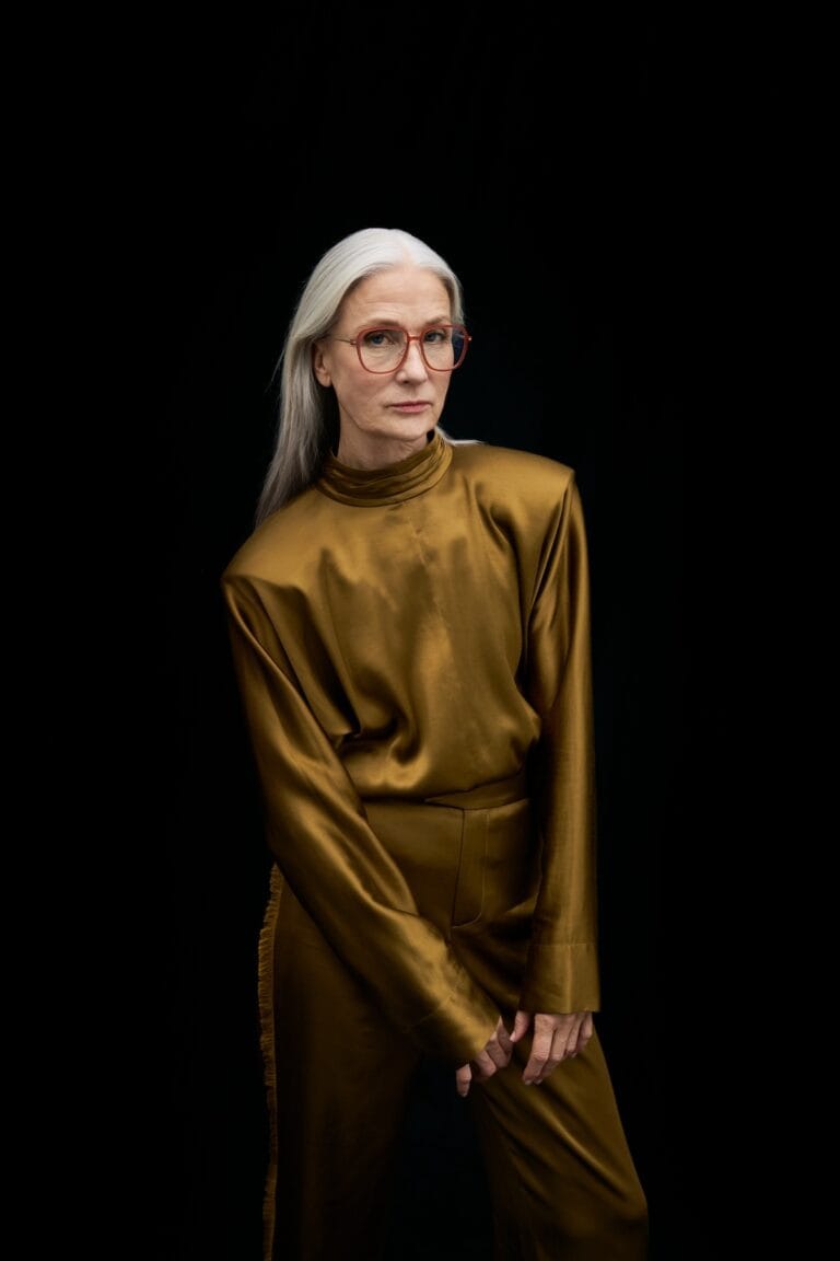 Portrait of an Older Woman with Gray Hair and Red Glasses Against a Black Background, Wearing a Gold Satin Blouse.  | Eyestylist 27th April 2026