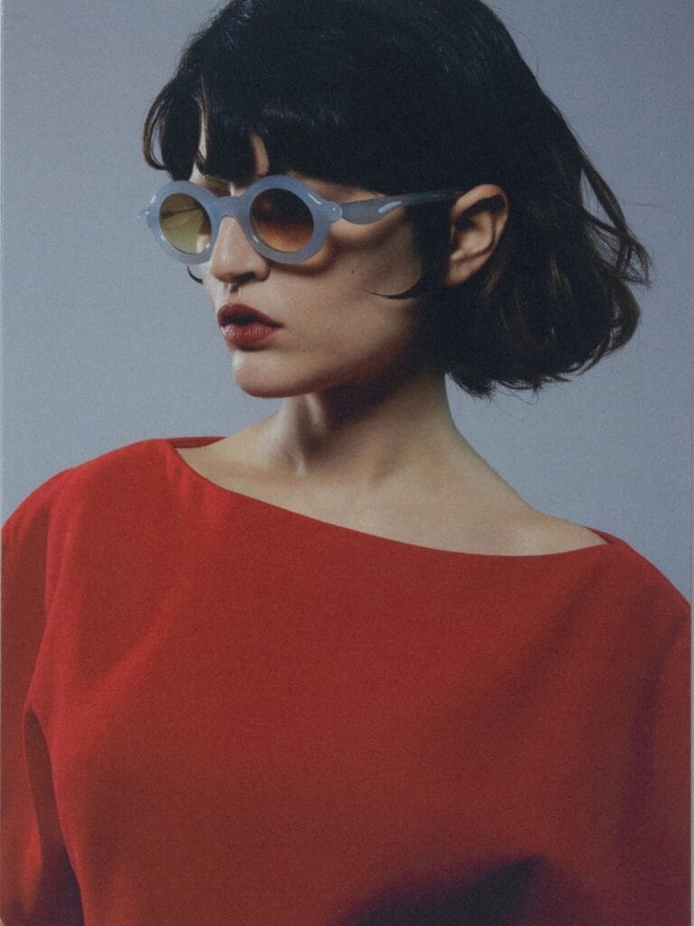 Woman with Short Black Hair Wearing Blue Round Sunglasses and a Red Boat-neck Top Against a Gray Backdrop.  | Eyestylist 20th April 2026