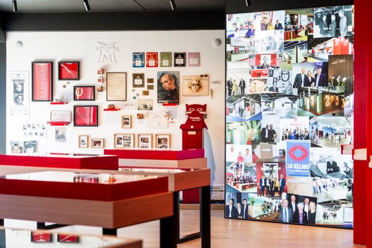 Exhibit Wall with Certificates, Photos, and a Red Sculpture, Set Beside Wooden Tables in a Gallery-like Room.  | Eyestylist 11th May 2026