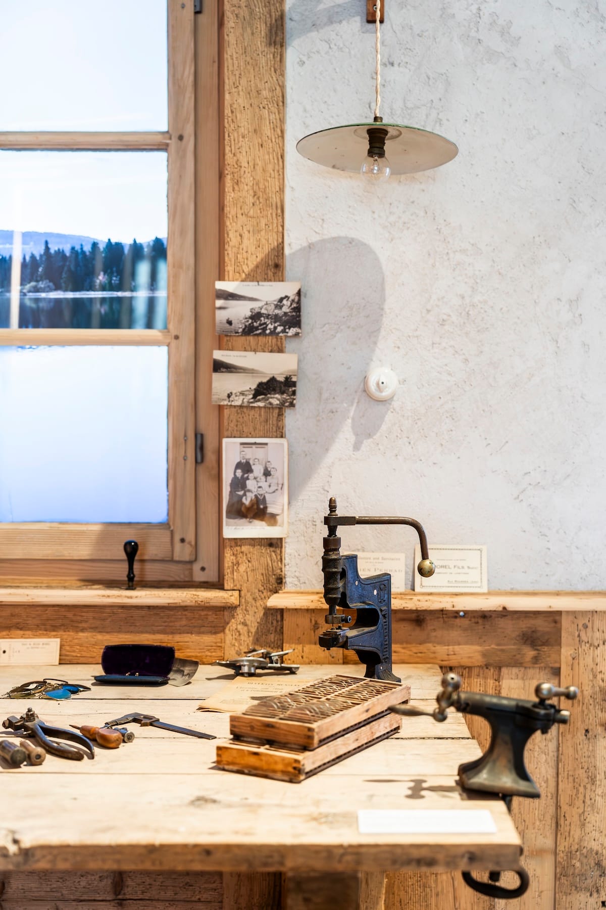 Vintage workshop scene: wooden workbench with scattered hand tools, a blue metal press, and black-and-white photos pinned to a beam by a window.