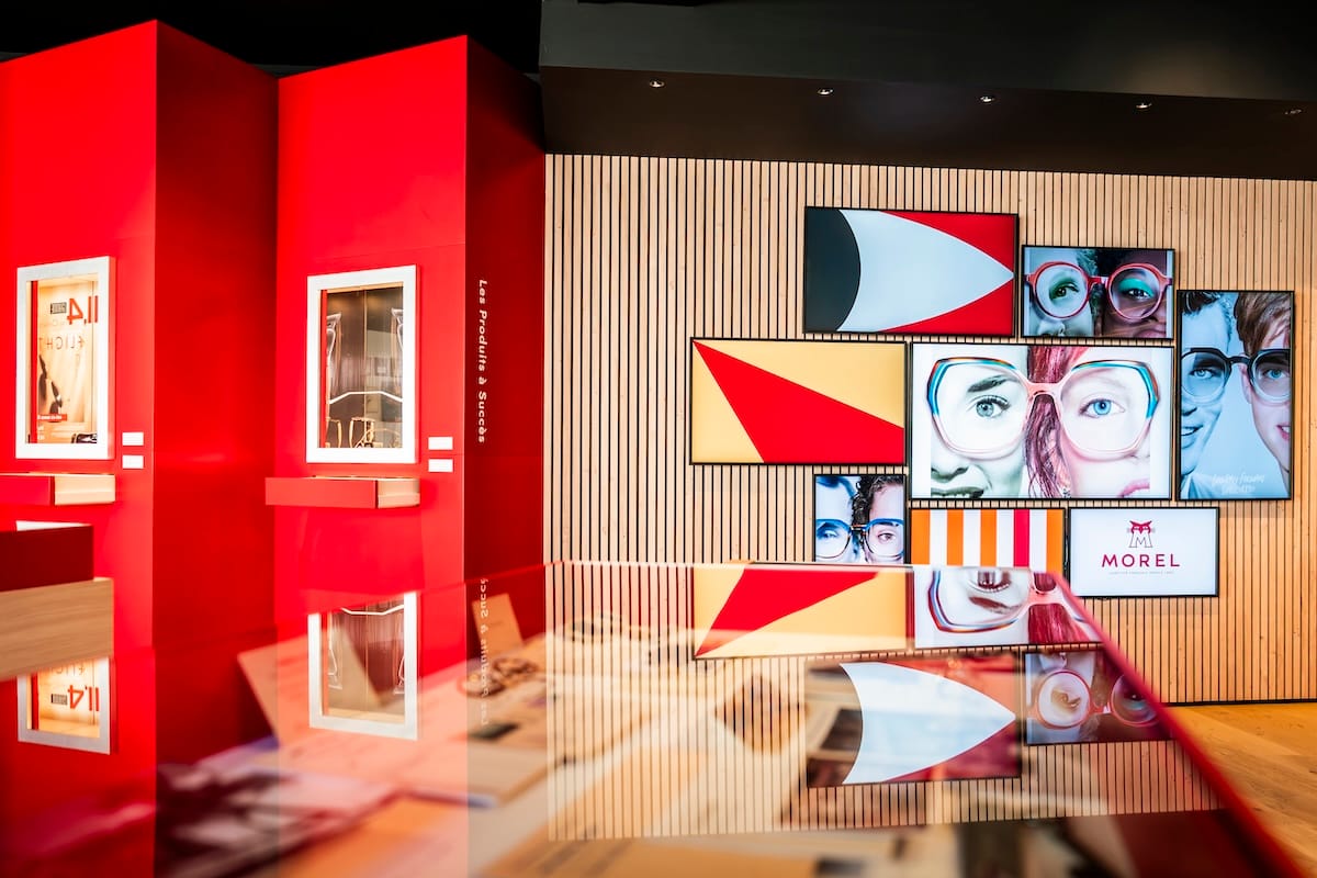Modern showroom with red walls, white-framed display cases, and a collage of colorful screens on a wood-paneled wall.