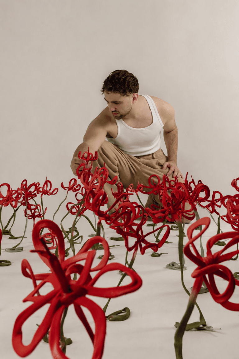 520629D1-ED17-47C6-8838-C493701F9D57 | Eyestylist Man in a White Tank Top Crouches Among Tall Red Curling Flower Sculptures in a Neutral Studio. | Eyestylist 26th May 2026