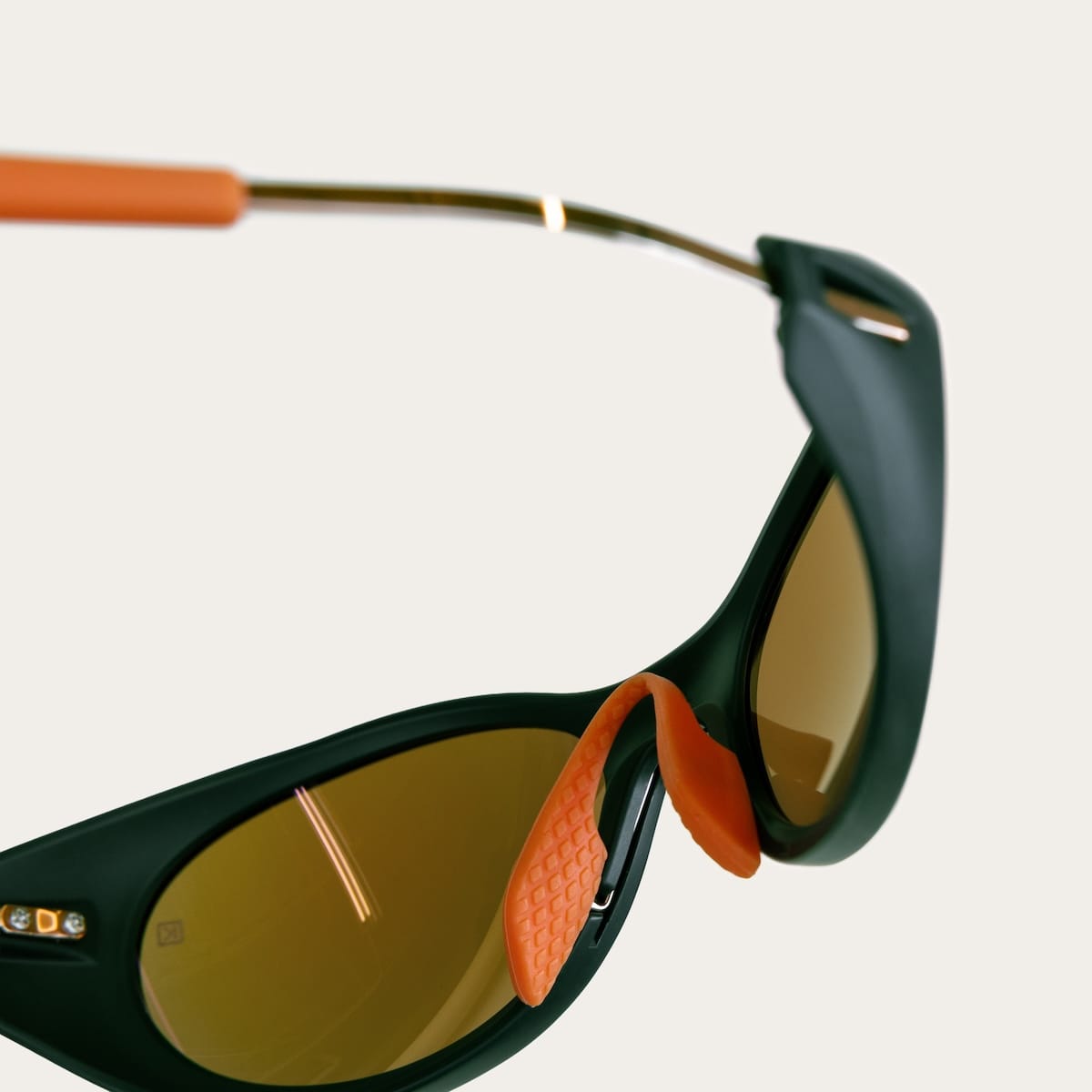 Close-up of dark green sunglasses with brown lenses and orange, textured nose pads; hinge and temple detail visible.