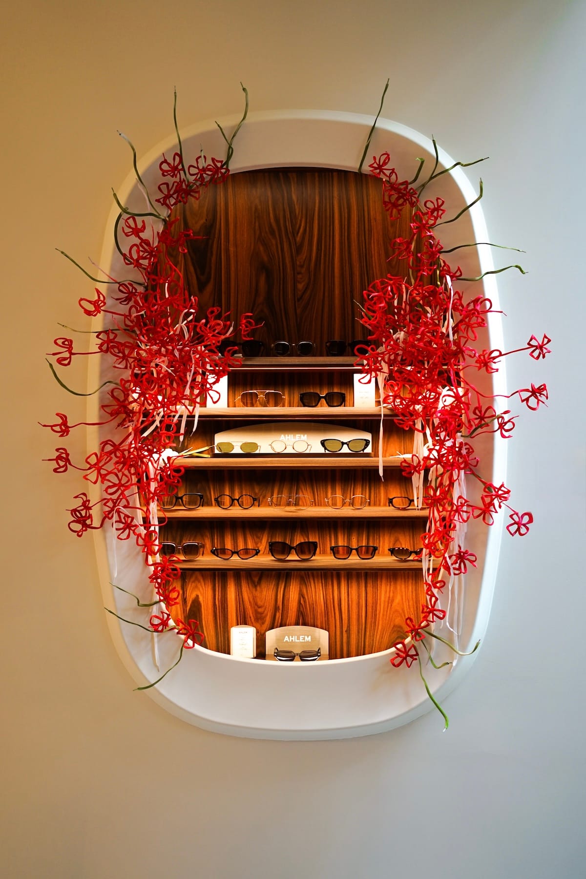 Oval white display case with wooden shelves holding rows of sunglasses, framed by red decorative flower garlands.