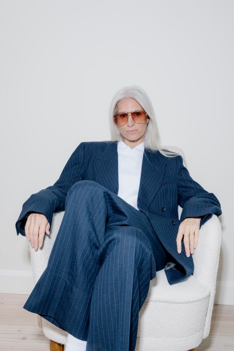 Person with Long White Hair Wearing a Blue Pinstripe Suit and Brown-tinted Glasses Sits in a White Chair, Looking at the Camera.  | Eyestylist 29th June 2026