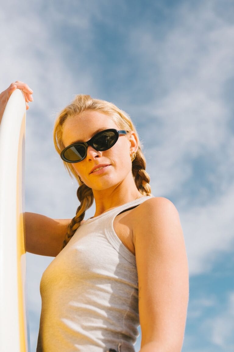 BIRDIE44 | Eyestylist Woman with Braided Hair and Sunglasses Rests Against a Surfboard Under a Blue Sky, Ready to Surf. | Eyestylist 18th June 2026