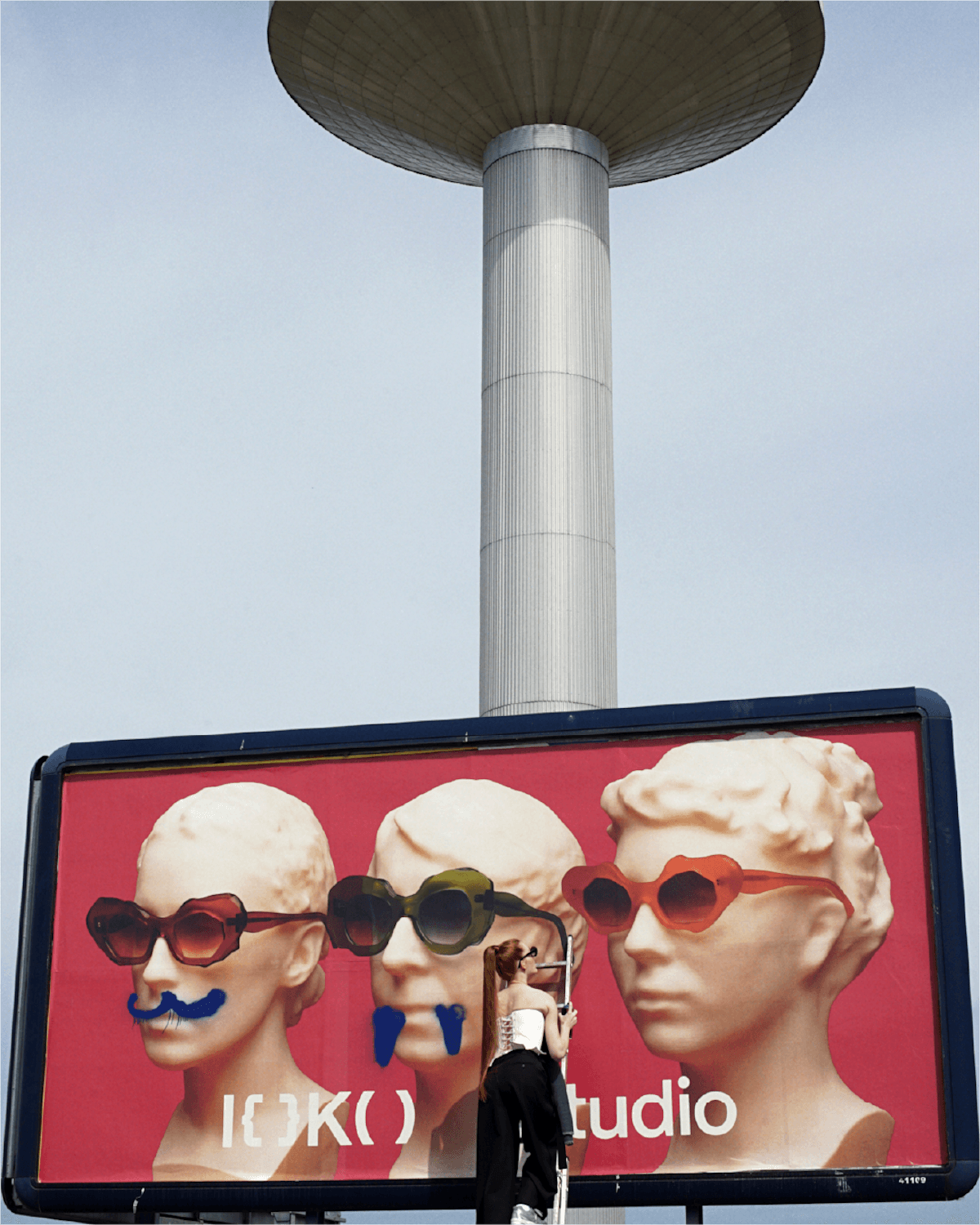 Person on a ladder adjusting a pink billboard showing three oversized heads wearing large sunglasses, with a tall cylindrical tower in the sky behind.