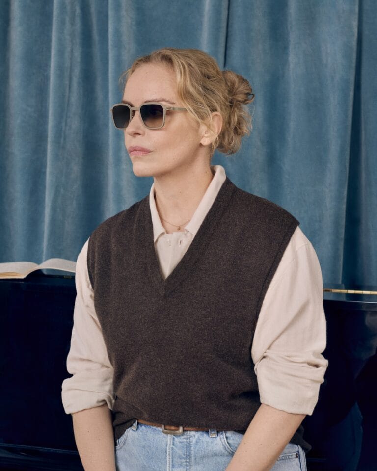 Person Wearing Sunglasses and a Brown Vest over a Light Shirt, Seated Beside a Piano with a Blue Curtain Backdrop.  | Eyestylist 5th June 2026