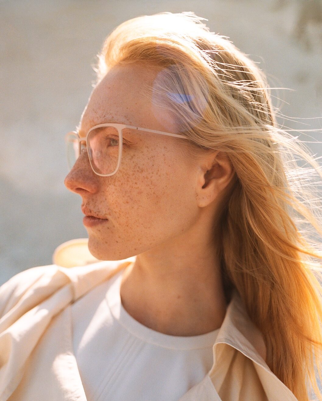 Side profile of a blonde woman wearing large rectangular glasses, freckles across her face, and a light beige jacket in sunny outdoor light.