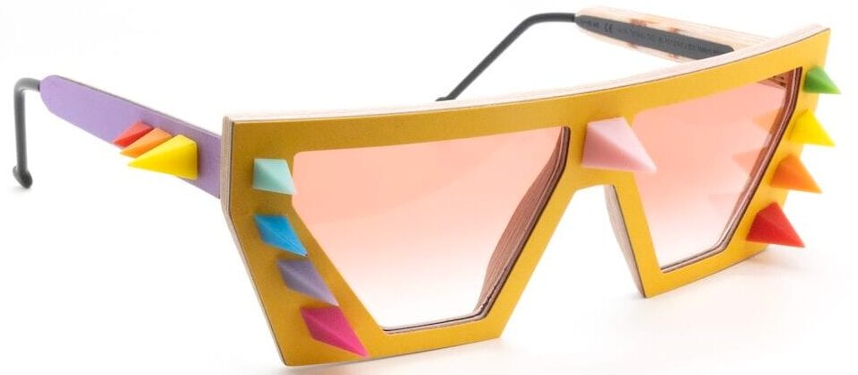 Colorful novelty sunglasses with a yellow frame, pink tinted lenses, and rainbow spikes along the sides and front.