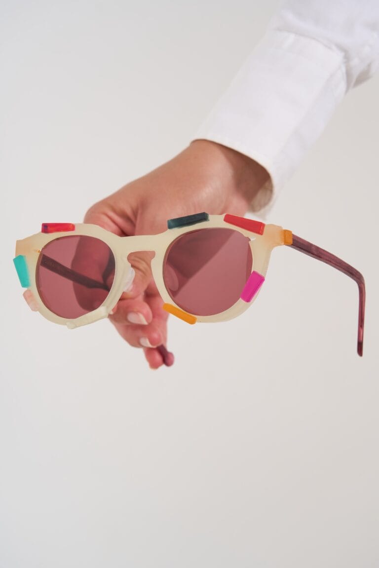 Hand Holding a Pair of White-framed Sunglasses with Pink Lenses and Colorful Clip-on Accents on the Frame Set Against a Plain Background.  | Eyestylist 16th July 2026