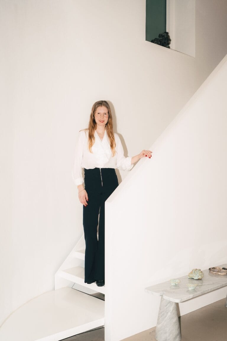 J2 copy | Eyestylist Woman with Long Blond Hair and Glasses Standing on a White Staircase, Wearing a White Blouse and Black Pants. | Eyestylist 6th July 2026