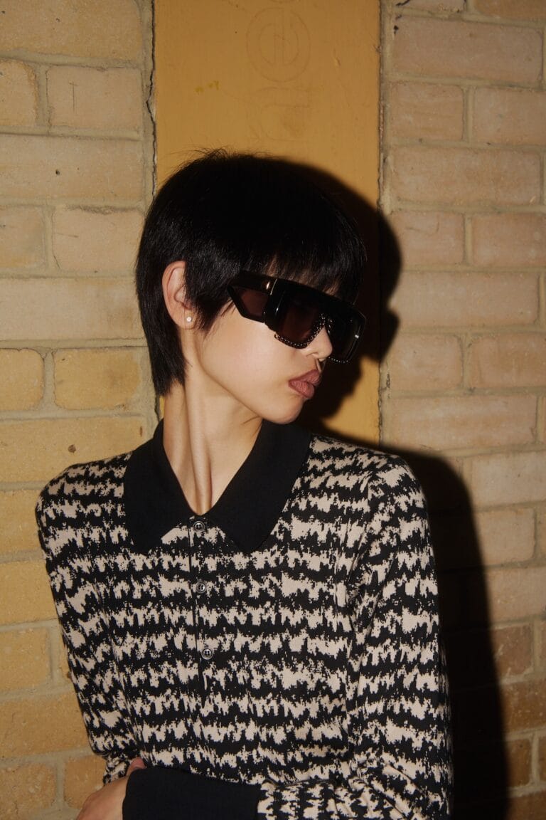 Person with Short Dark Hair Wearing Oversized Black Sunglasses and a Black-and-white Houndstooth Sweater, Posed Against a Brick Wall.  | Eyestylist 27th July 2026