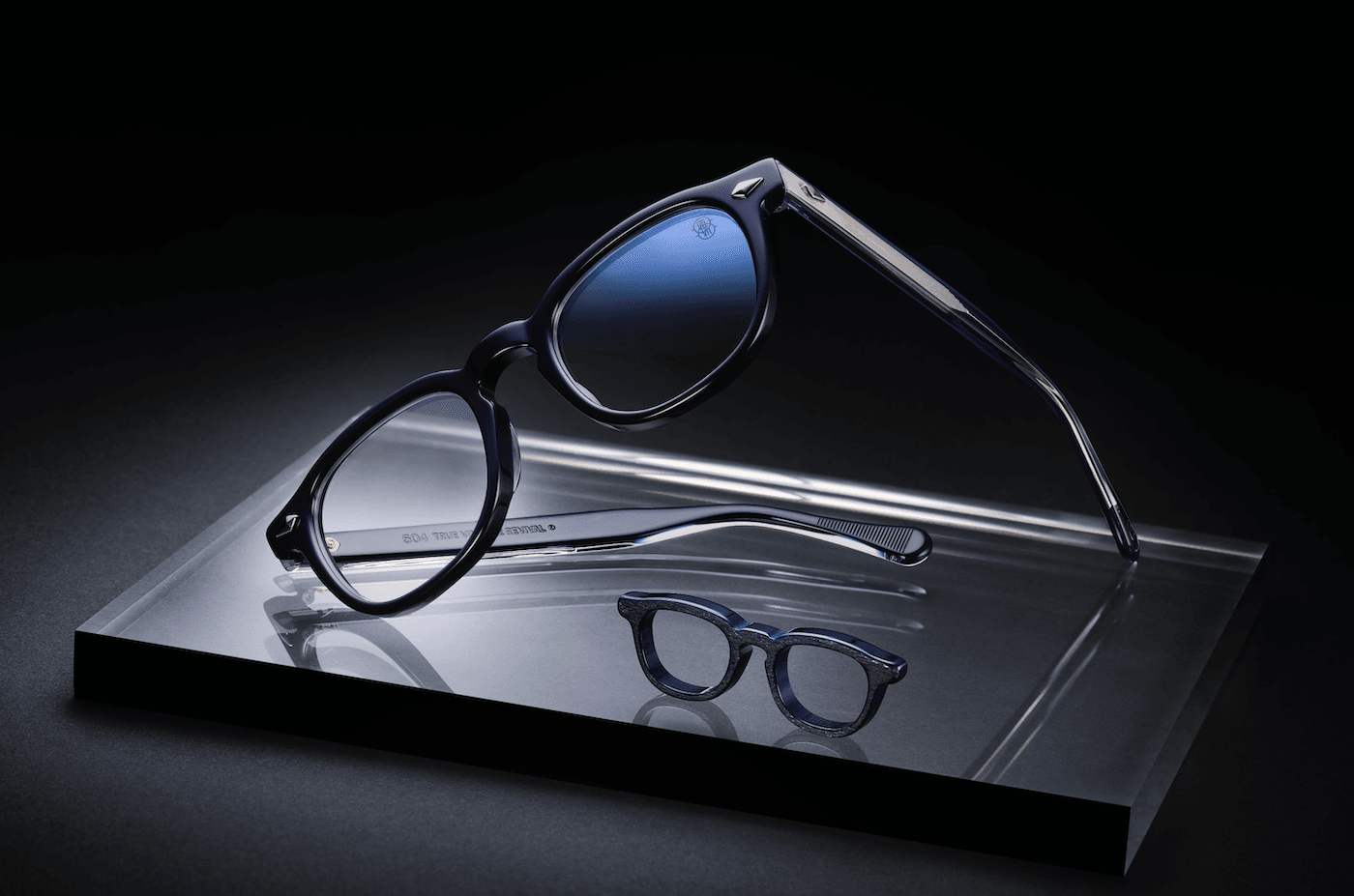 Two pairs of eyewear rest on a reflective glass display: a large dark-framed pair with blue-tinted lenses and a smaller round-frame pair in front, set against a black background.”,