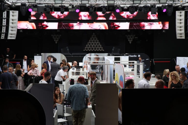 Crowded Convention Floor with Exhibitor Booths, a Black Stage, and a Long Led Video Wall Showing Fashion Portraits Above the Crowd.  | Eyestylist 24th August 2026