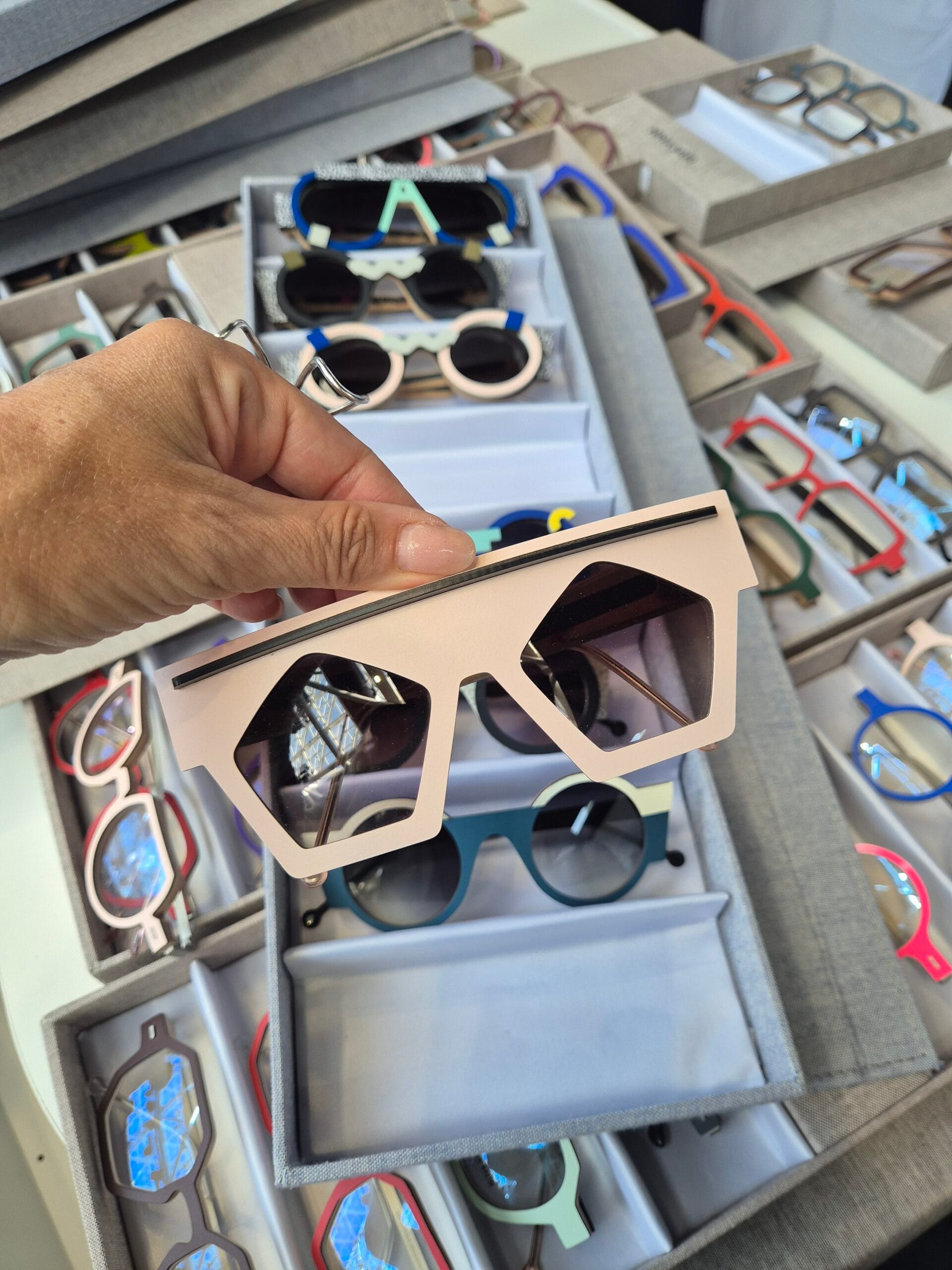 Hand holding pale pink square-frame sunglasses with dark lenses, among display boxes of colorful eyewears in a store