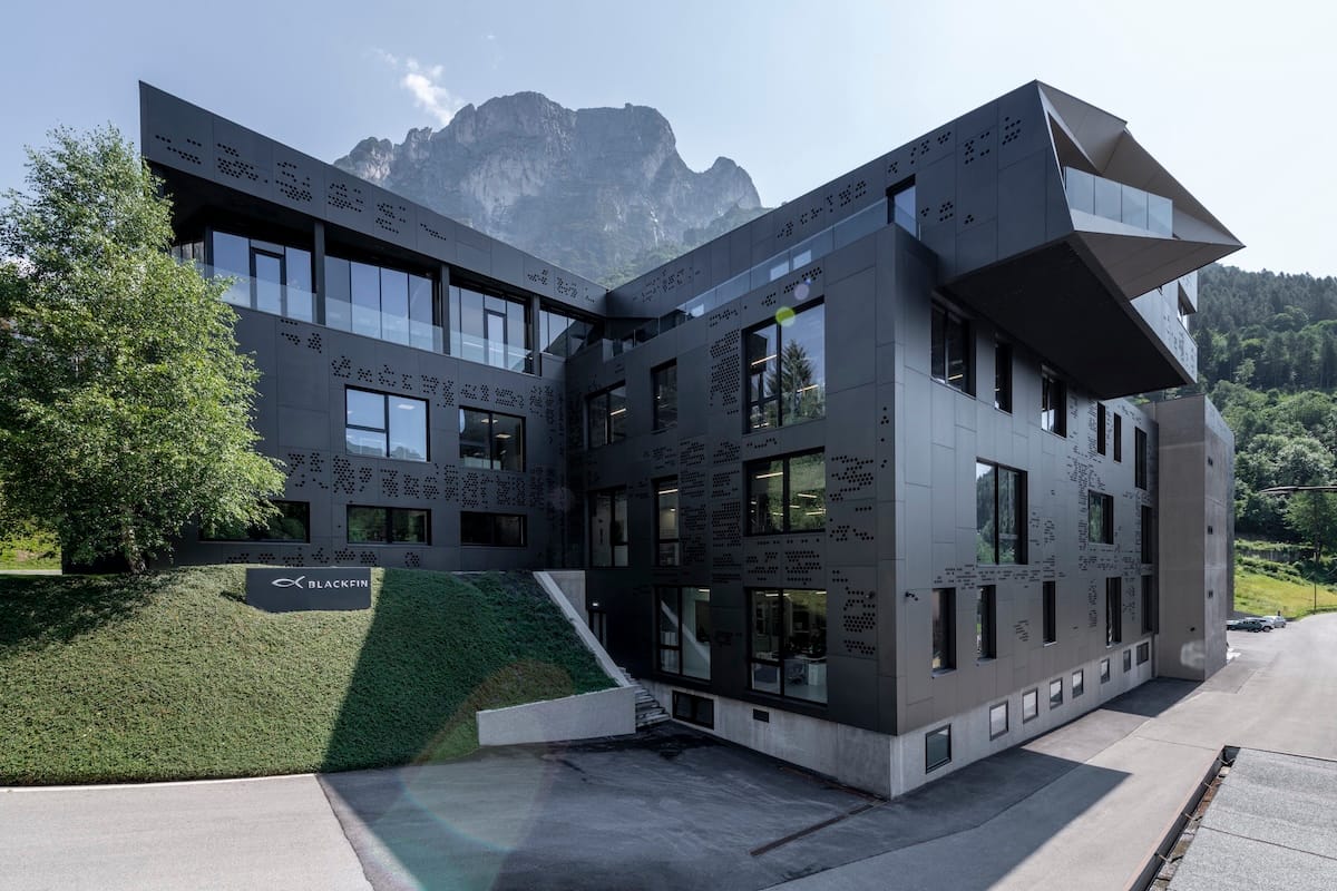 Modern dark gray office building with perforated panels, large glass windows, and a Blackfin sign on a grassy slope with alpine mountains in the background.