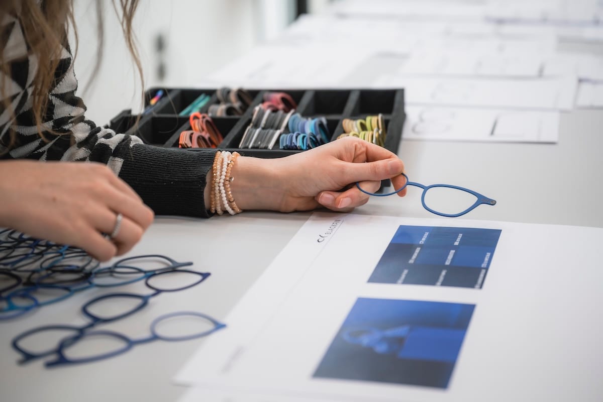 Person selecting a blue eyeglass frame over a white table with a color tray in the background