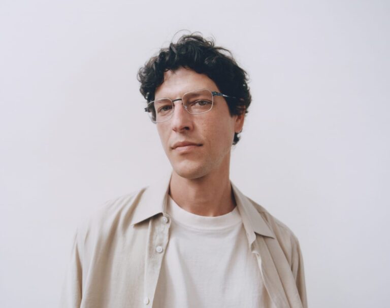 Head-and-shoulders Portrait of a Man with Curly Dark Hair and Glasses, Wearing a Beige Shirt over a White Tee, Against a Plain Light Background.  | Eyestylist 12th August 2026
